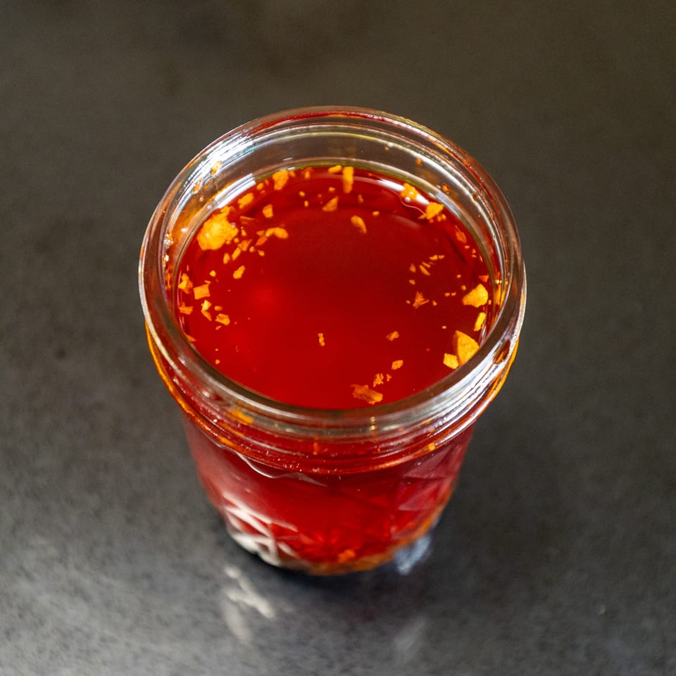 A glass jar of vibrant red Thai chili oil with golden flakes, viewed from above.