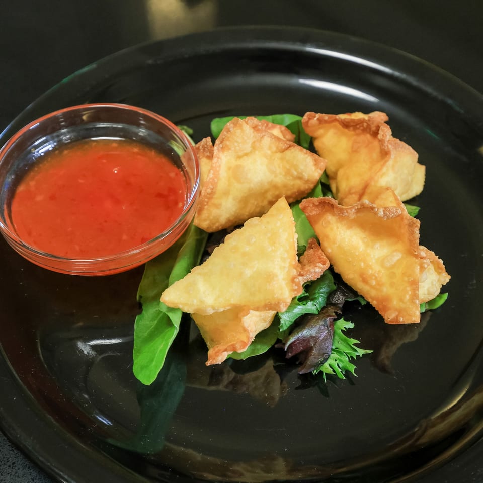 Golden-fried Thai Esane wontons with a side of sweet chili dipping sauce on a black plate.