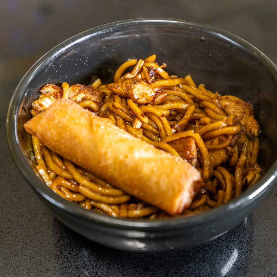 Thai Esane's savory noodles with chicken and a crispy fried spring roll in a black bowl.